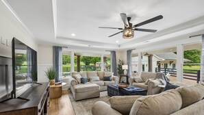 A spacious living room with a flat-screen TV, comfortable seating, a ceiling fan, and large windows offering a view of the outdoors.