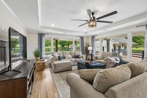 A spacious living room with a flat-screen TV, comfortable seating, a ceiling fan, and large windows offering a view of the outdoors. - Your Franklin Oasis (Franklin)