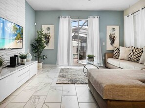 Ein modernes Wohnzimmer mit einer Couch, einem Couchtisch und einem Fernseher auf einem weißen Schrank. An den Wänden befinden sich Pflanzen und Kunstwerke.