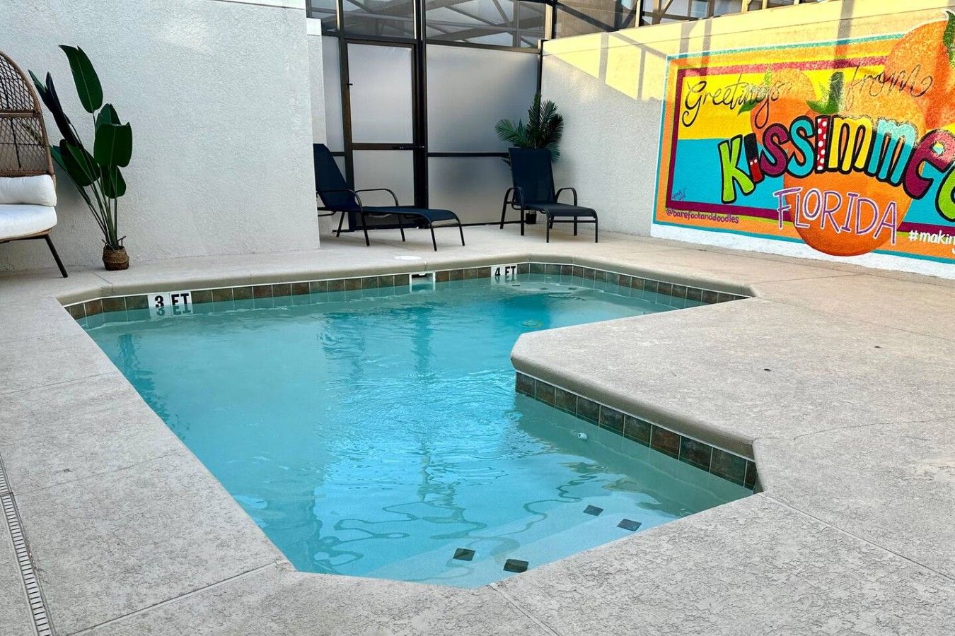 A swimming pool with a tiled edge, a lounge area with chairs, and a wall with a colorful mural.