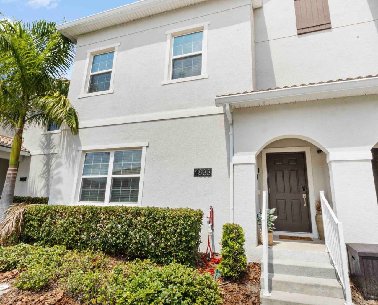 A two-story residential building with a white exterior, a dark front door, and a small front yard with a palm tree and shrubs.