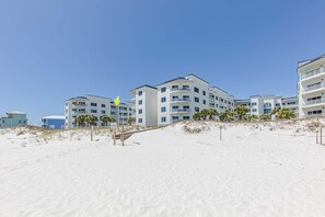 A beachfront hotel with multiple balconies, a sandy beach in the foreground, and a clear blue sky. - Read  the Reviews!Right on the Beach with East and West Views! Cannot get any closer to the Water! (Orange Beach)