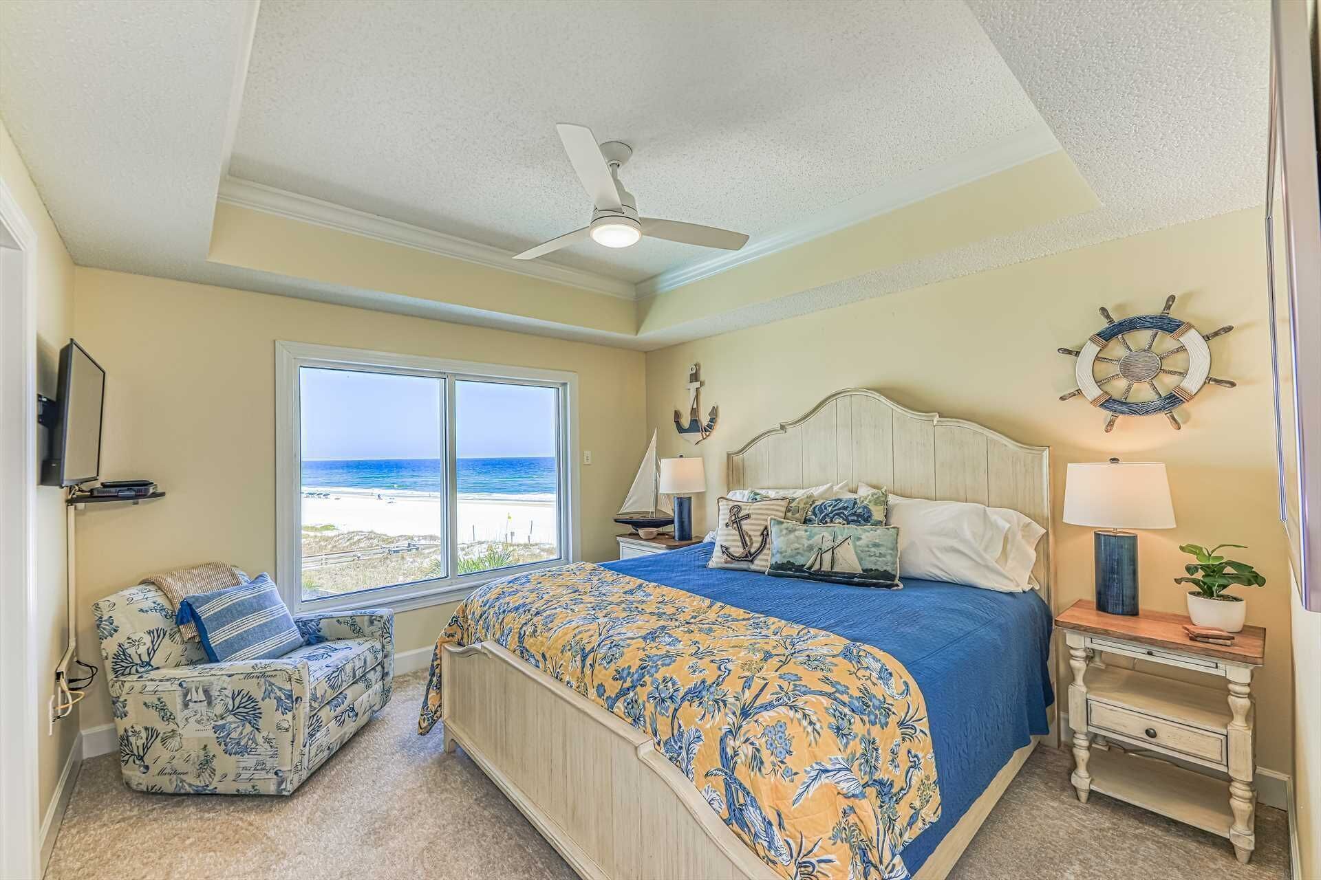 A bedroom with a large bed, a chair, a desk, a ceiling fan, and a view of the beach.