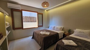 Une chambre d’hôtel avec deux lits, une fenêtre avec des stores, une table de chevet avec une plante et un climatiseur fixé au mur.