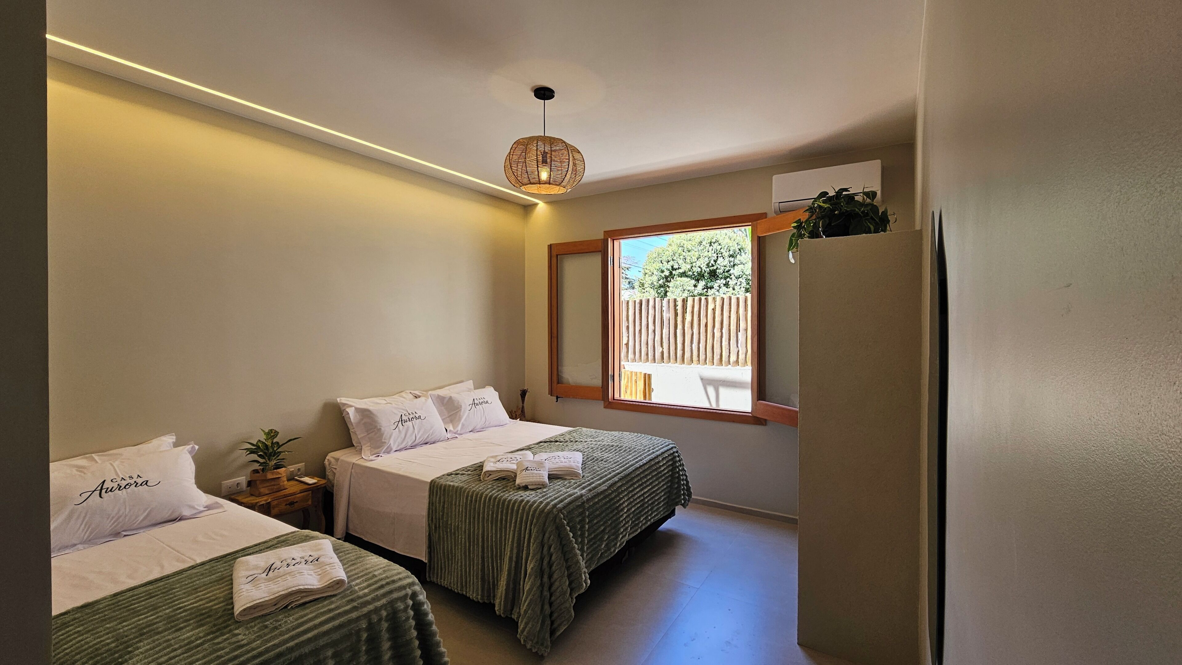 A hotel room with two beds, a window, a hanging light fixture, and a small plant.