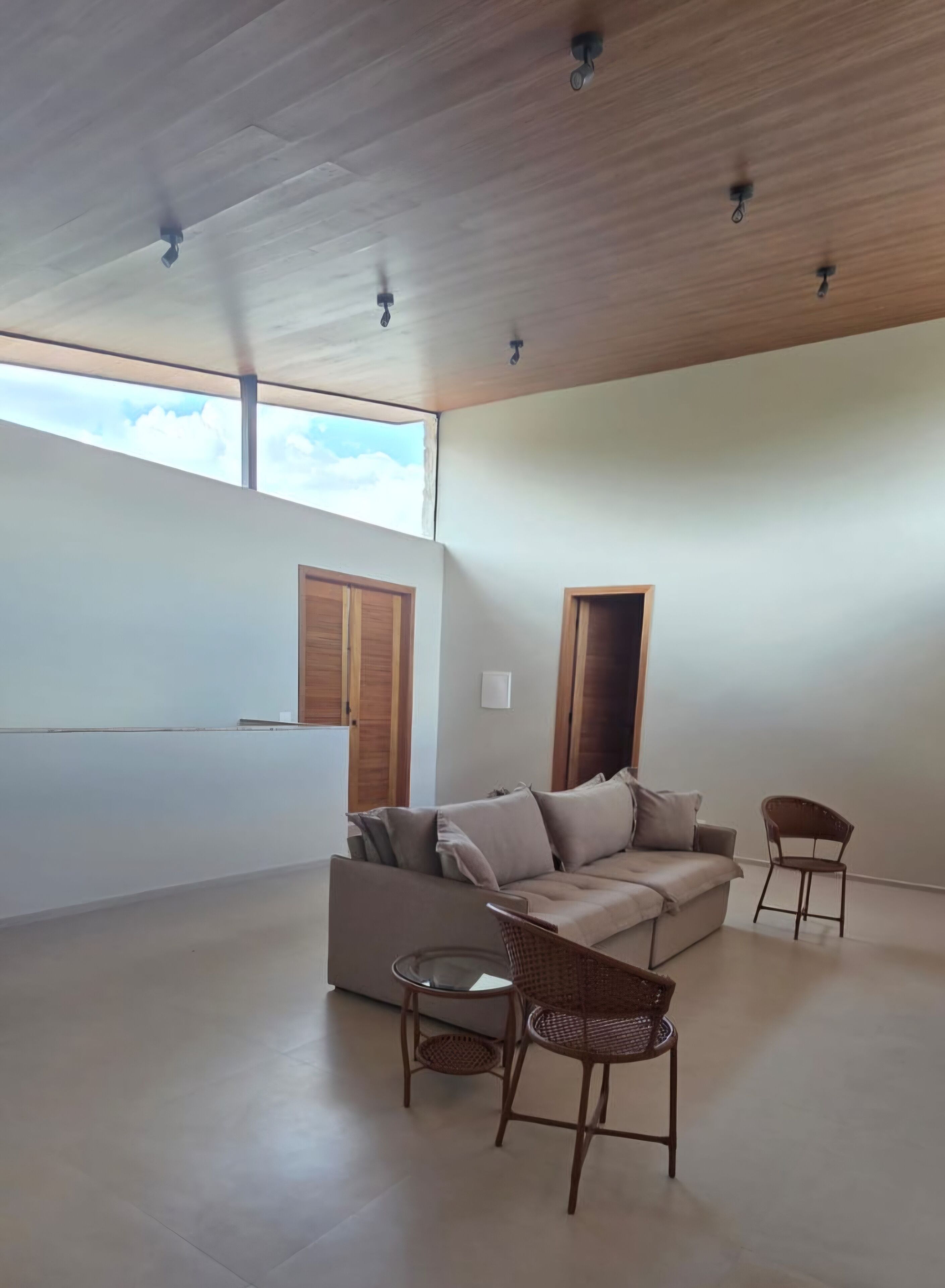 A spacious living area with a high ceiling, a sofa, two wicker chairs, and a window with wooden shutters.