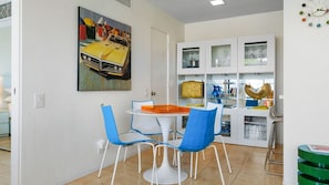 A dining area with a round table and blue chairs, a kitchen with cabinets and a sink, and a wall with a painting of a car. - Whimsical mid-century mod at famed Ocotillo Lodge (Palm Springs)