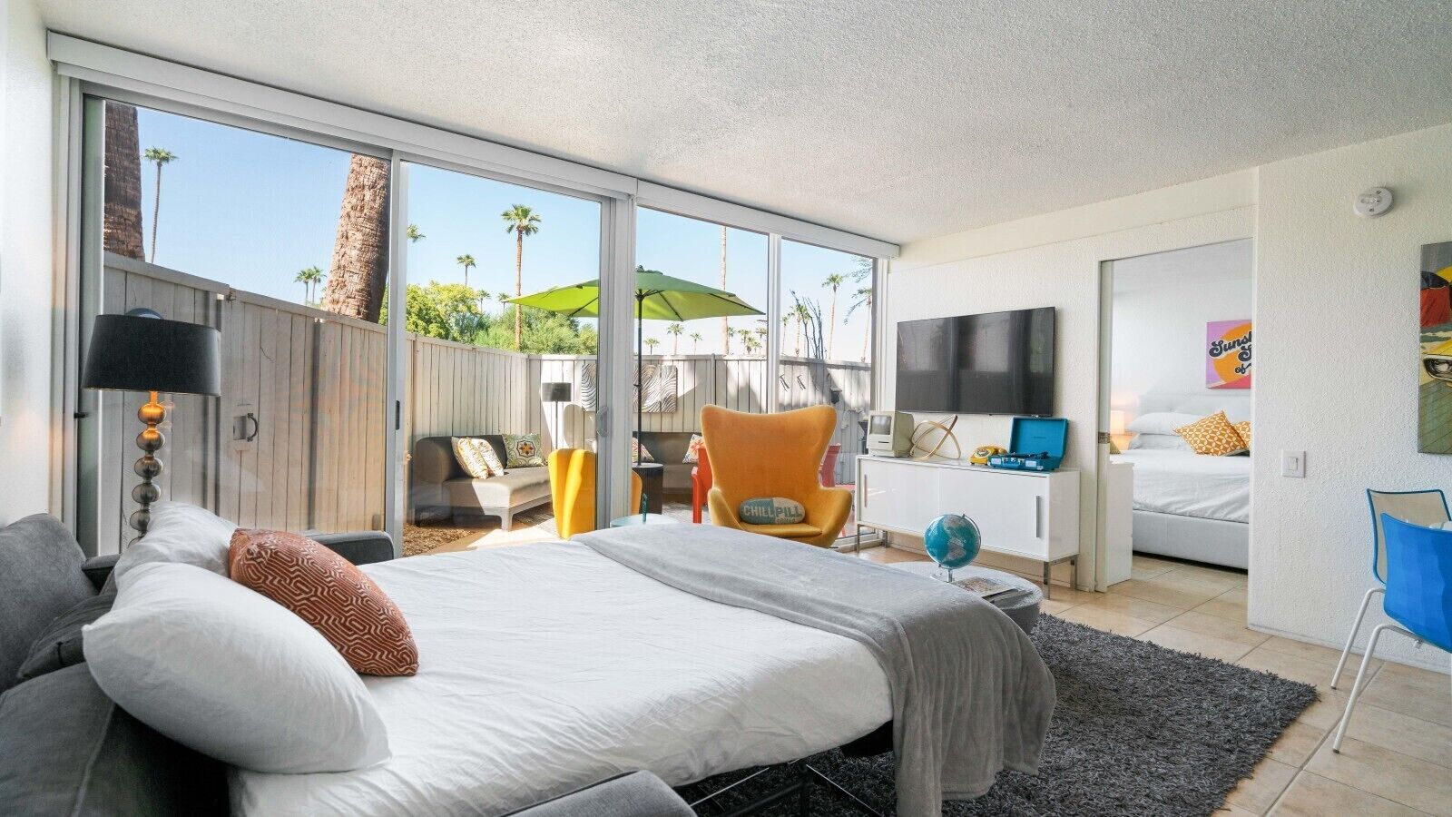 A modern bedroom with a large bed, a sofa, a chair, a TV, and a view of palm trees.
