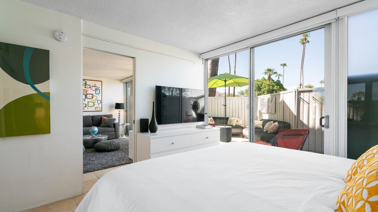 Ein Schlafzimmer mit einem großen Bett, einem Fernseher, einer Schiebetür zu einem Balkon und Blick auf Palmen.