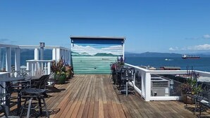 Una terrassa de fusta amb seients exteriors, una zona coberta amb un mural, i vista al mar i vaixells.