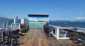 Een houten terras met buitenmeubilair, een overdekt gedeelte met een muurschildering en uitzicht op zee en schepen.