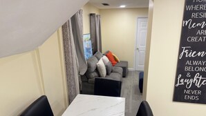 Uma área de refeições com mesa de mármore branco e cadeiras pretas, um sofá cinza com almofadas, e uma parede com uma frase motivacional.