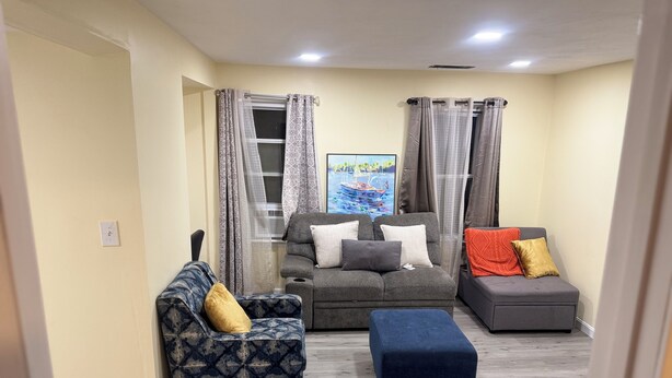 Sala de estar com sofá, uma poltrona e um banquinho. Há uma janela com cortinas, um quadro na parede e uma luminária no teto.