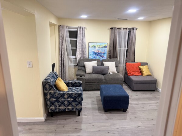 A living room with a sofa, a chair, and an ottoman. There is a window with curtains, a painting on the wall, and a ceiling light. - Delightful 3-bedroom apartment with AC in charming Johnston (Johnston)