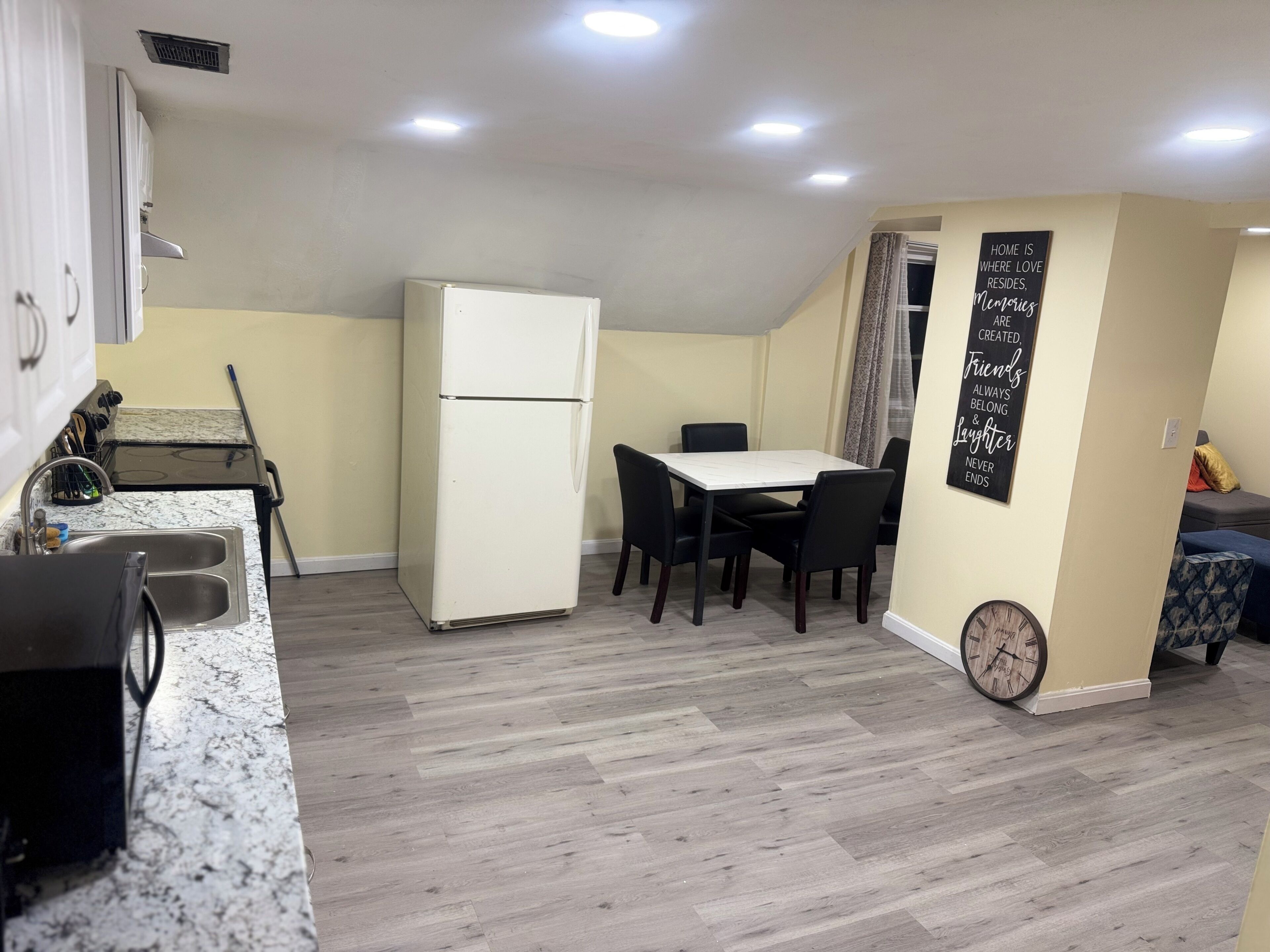 A compact kitchen with a refrigerator, a dining table with chairs, and a clock on the wall.