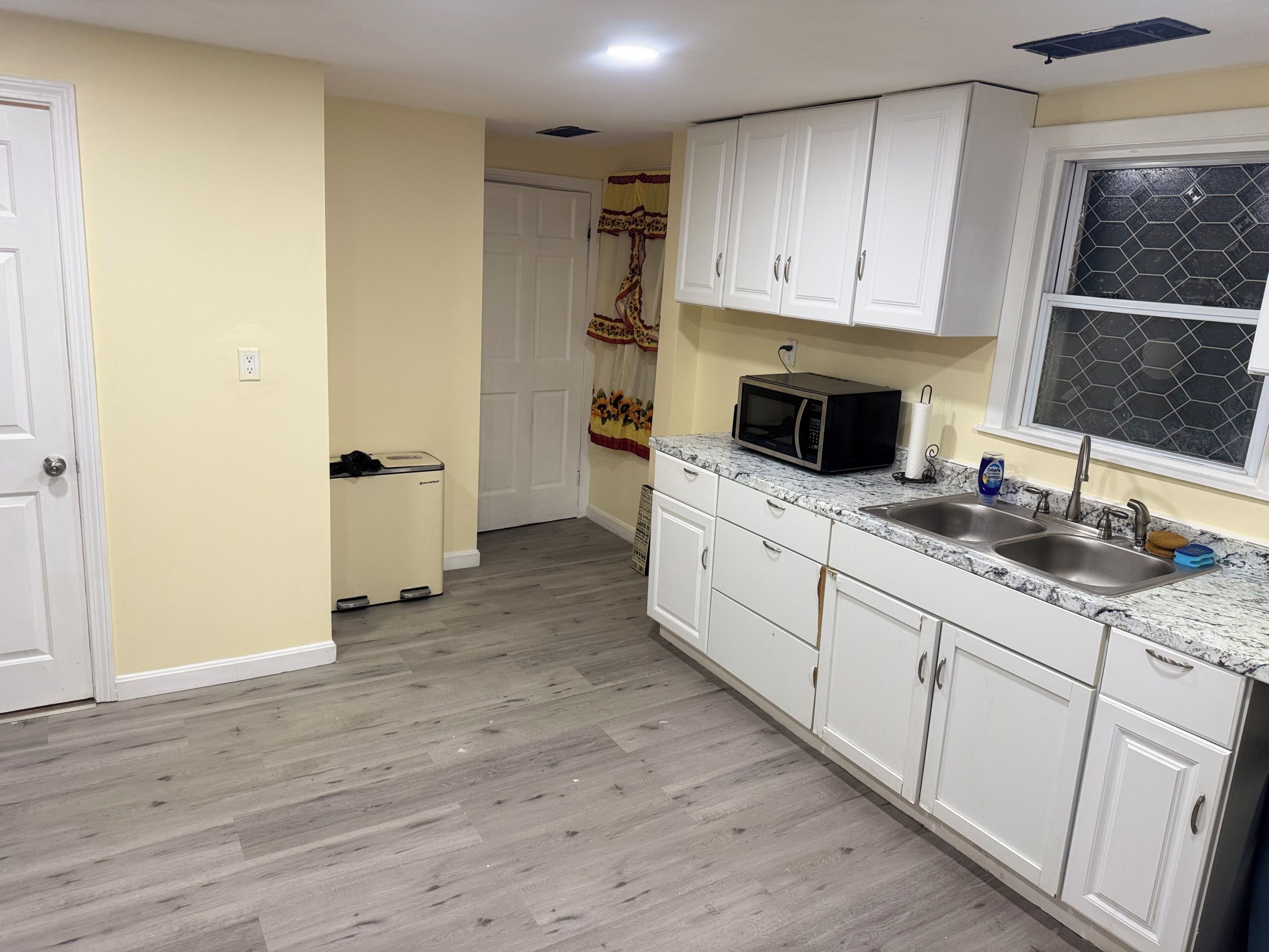 A kitchen with white cabinets, a microwave, a sink, and a window with a hexagonal pattern.