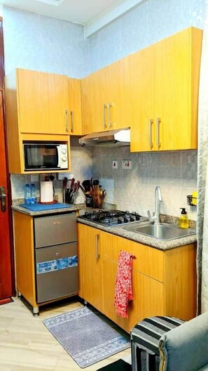 A compact kitchen with wooden cabinets, a microwave, a gas stove, and a sink. - COZY NEST Apt| Wi-Fi | Near town| Laundry Rm | Generator (Dar es Salaam)