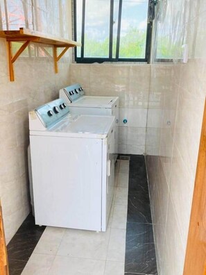 A compact laundry room with a washing machine and dryer, a window with a view, and a wooden shelf. - 7 - Min Walk to Beach | Pool - KING Bed | Wi-Fi (Jambiani)