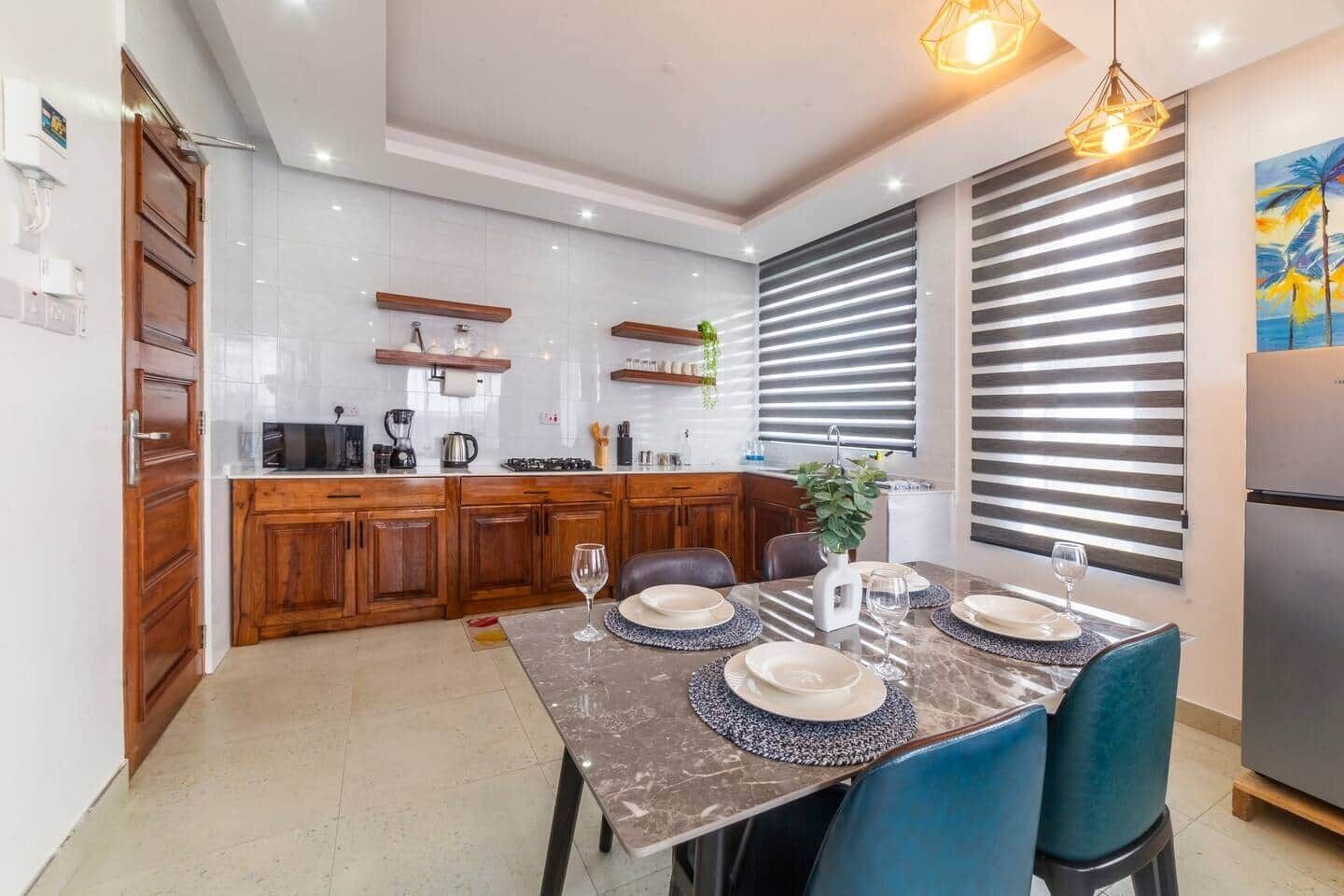 A modern kitchen with wooden cabinets, a dining table set for four, and a large window with blinds.