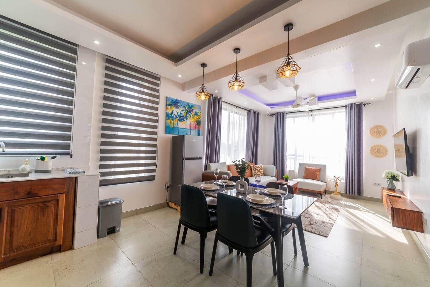 A modern living room with a dining area, a kitchen, and a large window with blinds.