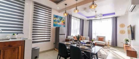 A modern living room with a dining area, a kitchen, and a large window with blinds.