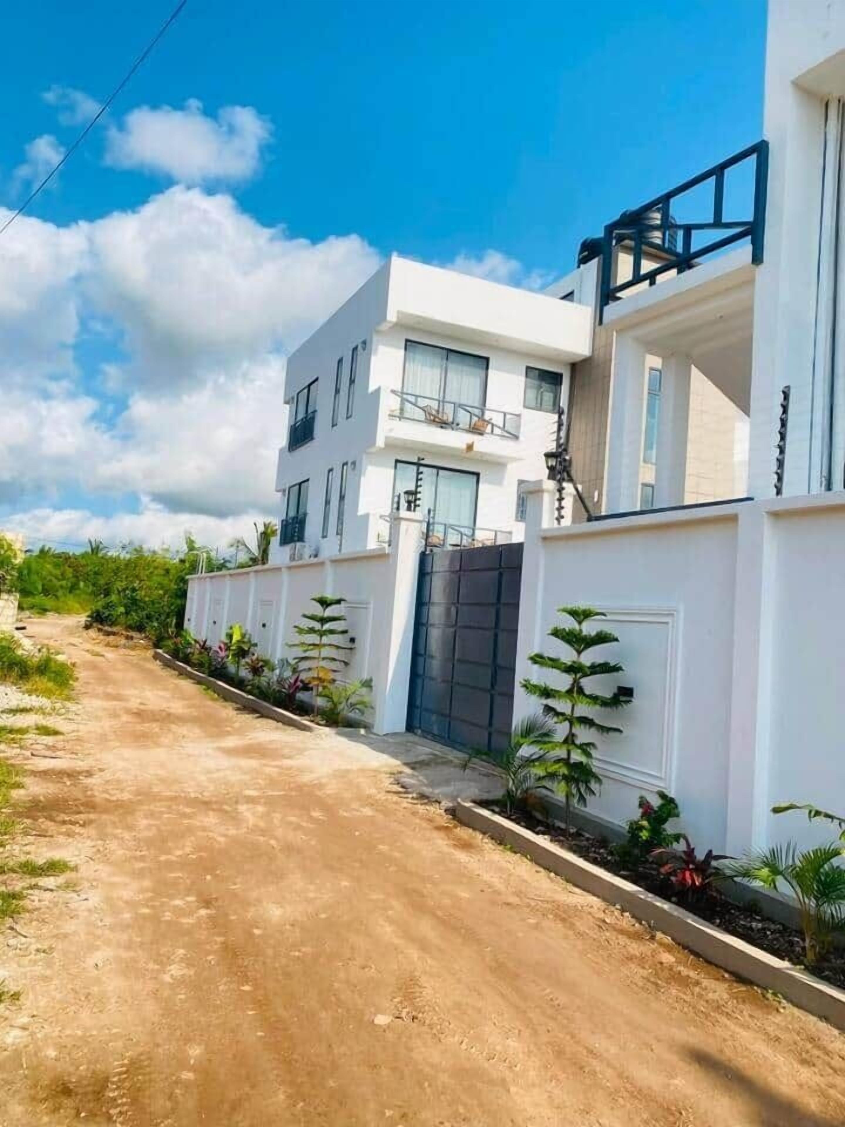 Un edificio residencial moderno de dos pisos con fachada blanca, entrada con reja y camino de acceso de tierra.