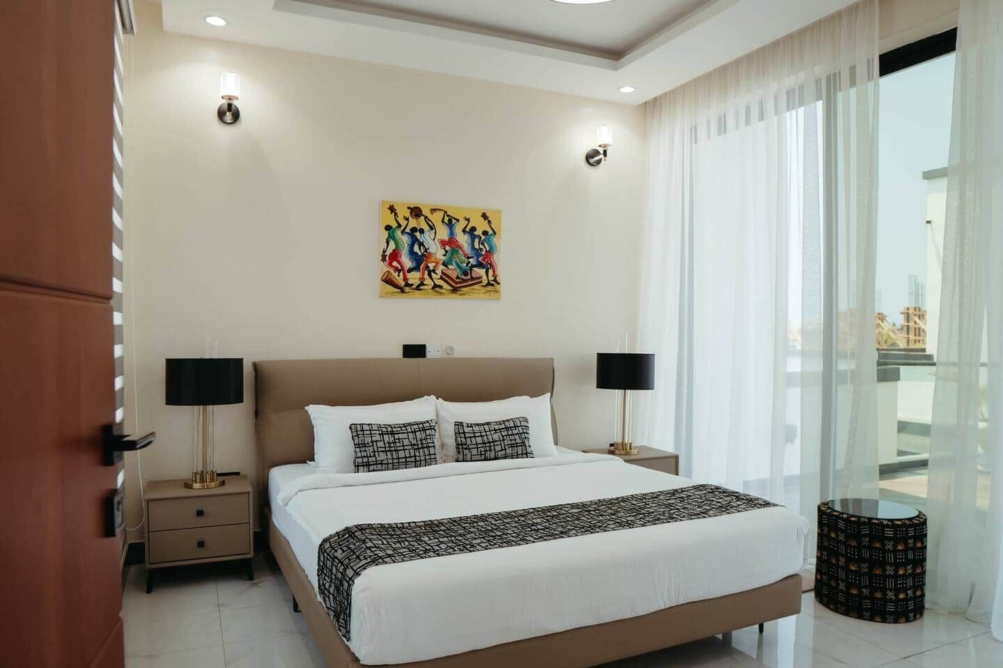 Une chambre d’hôtel avec un grand lit, deux tables de chevet, une œuvre d’art accrochée au mur et une vue à travers des rideaux transparents.
