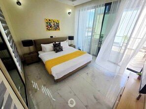 A hotel room with a bed, a nightstand, a lamp, a painting on the wall, and a view of the outdoors through large windows. - New Modern Bliss apt- 3Bdrm w/pool- gym-hottub (Dar es Salaam)