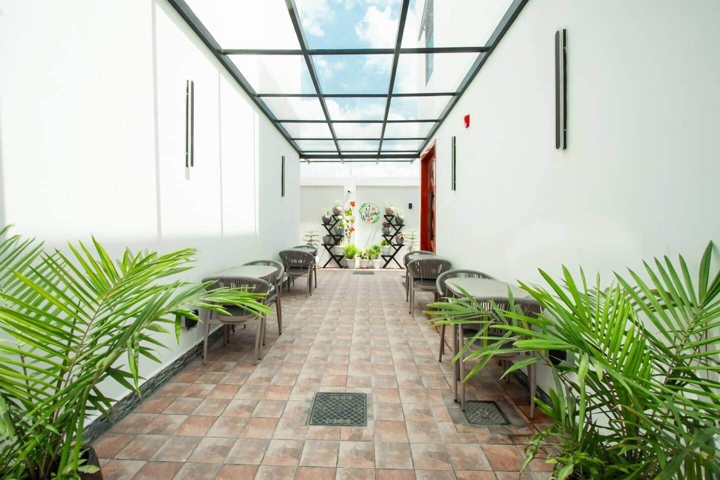 A covered outdoor seating area with chairs and tables, surrounded by potted plants, and a view of the sky through a glass ceiling.
