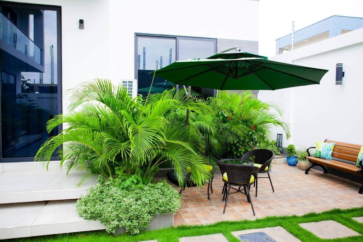 A modern outdoor patio with a green umbrella, wicker furniture, and lush greenery.