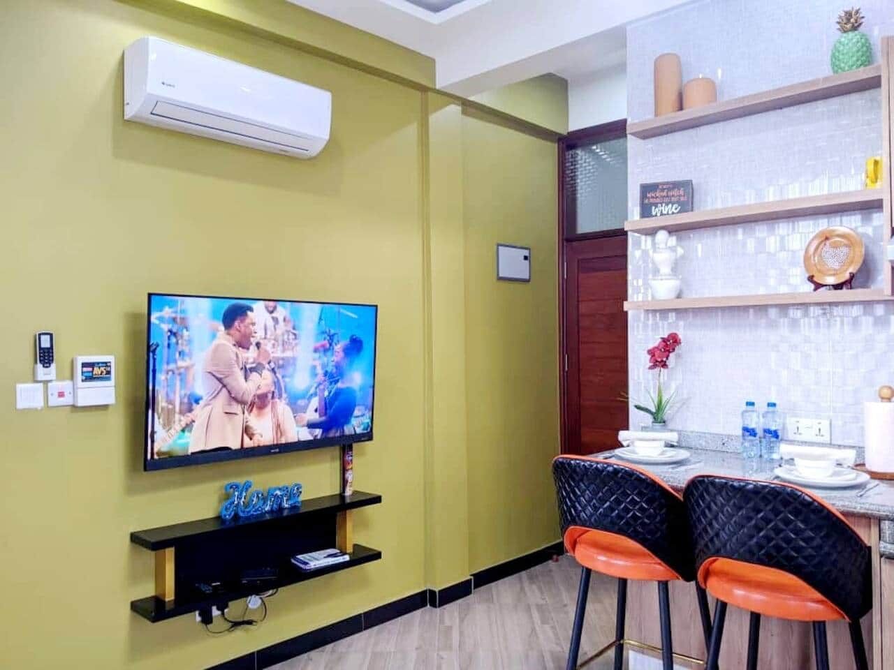 Uma sala de estar com televisão de tela plana, uma pequena estante com livros e um espaço de jantar com mesa e cadeiras.
