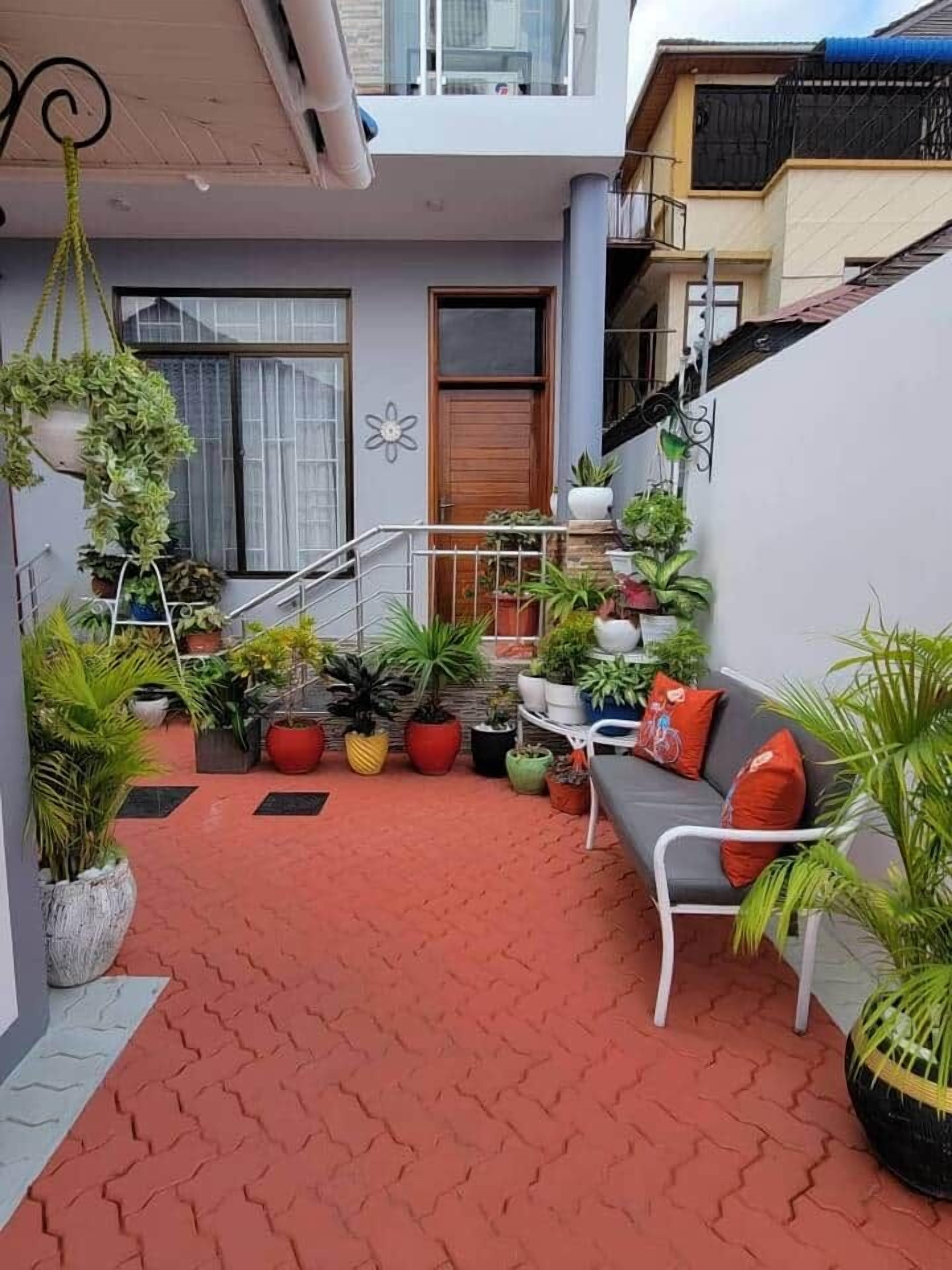 A covered outdoor area with a red tiled floor, a grey sofa with red cushions, and various potted plants.