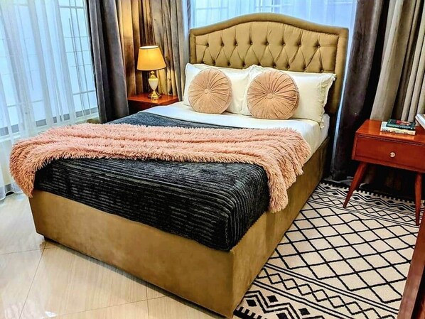 Un lit avec une tête de lit capitonnée, deux oreillers, une couverture moelleuse et un tapis à motifs.