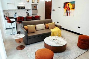 A modern living room with a sofa, two coffee tables, and a dining area with chairs and a kitchen in the background. - New Vibrant Charm w/Pool +HotTub | Gym | Wi-Fi (Dar es Salaam)