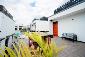 A modern rooftop terrace with a glass balustrade, a wooden table, a sofa, and a red door. - New Vibrant Charm w/Pool +HotTub | Gym | Wi-Fi (Dar es Salaam)