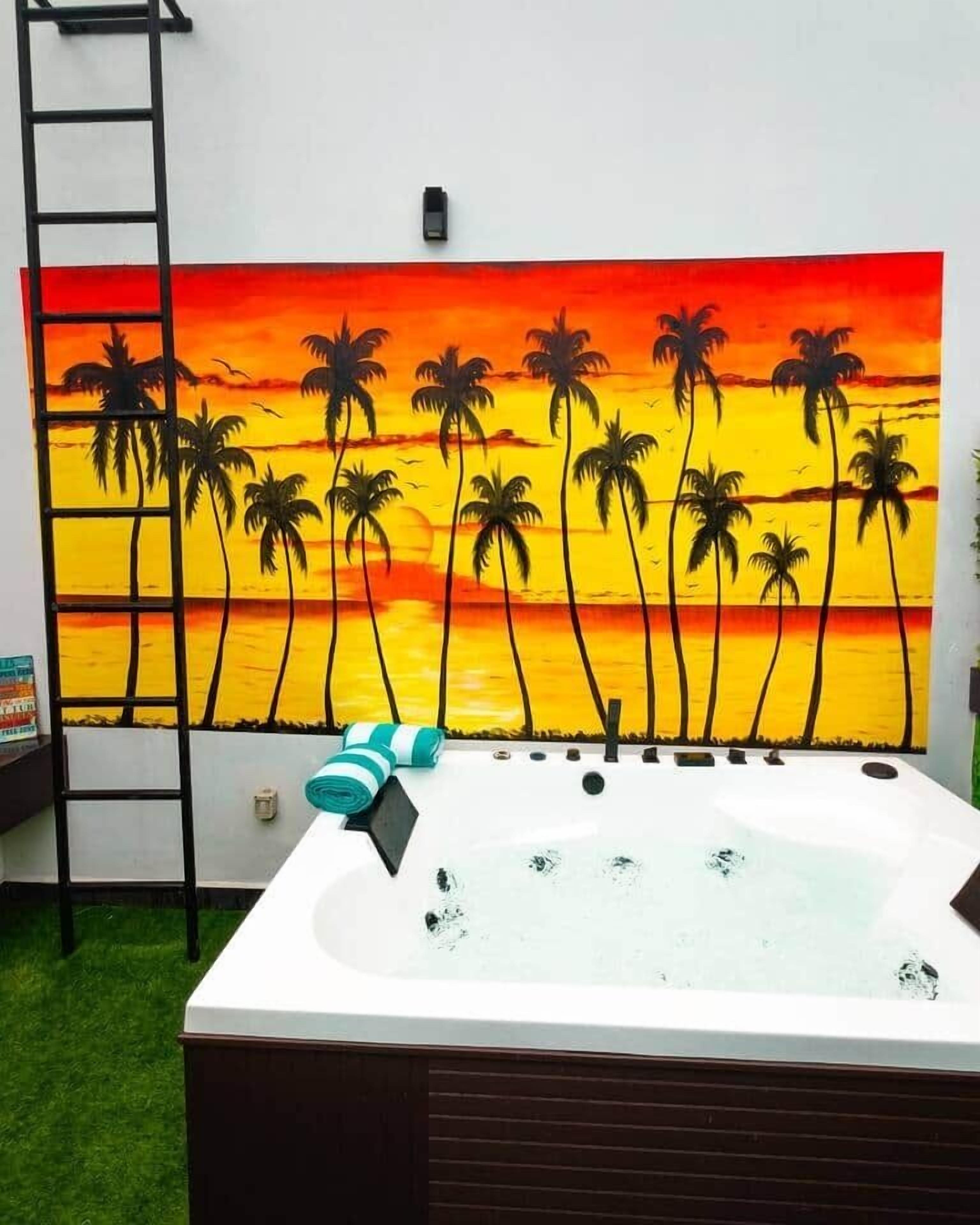 A room with a large painting of a sunset and palm trees on the wall, a hot tub, and a ladder.