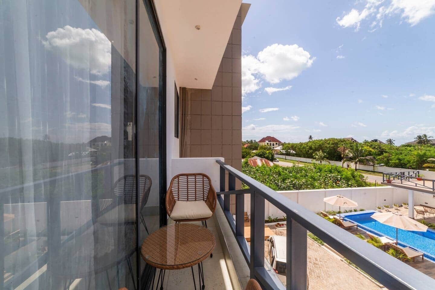 A balcony with two wicker chairs and a glass table, overlooking a swimming pool and greenery.