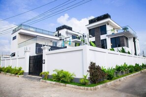 Una casa moderna de dos pisos con fachada blanca, un portón negro y un pequeño jardín con plantas tropicales.