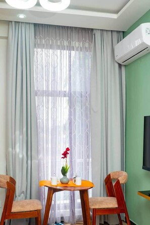 Ein Zimmer mit Tisch, zwei Stühlen, einem Fenster mit durchsichtigen Vorhängen und einer Klimaanlage an der Wand.