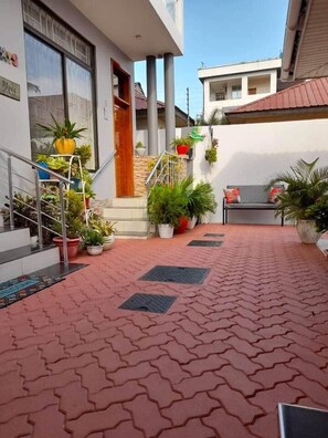 A paved courtyard with steps leading to a building entrance, surrounded by potted plants and a bench. - Serenity Peak Apt -King Bed - Kitchen-Laundry-Generator (Dar es Salaam)
