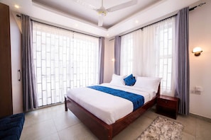 Une chambre avec un grand lit, une table de chevet, un ventilateur de plafond et une fenêtre avec des rideaux.