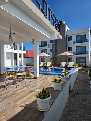 A rooftop pool area with lounge chairs and umbrellas, surrounded by a modern building with balconies. - Seashells Private Apt - Kitchen| Pool |Wi-Fi | Beachwalk (Jambiani Kikadini)