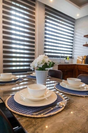 A dining area with a table set for four, featuring white plates and bowls, a vase with white flowers, and a view through large windows with horizontal blinds. - Ocean Breeze retreat - 7 min Beach walk | Pool (Jambiani Kikadini)