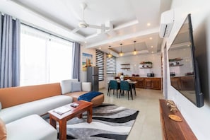 Uma sala de estar moderna com sofá, mesa de centro e área de jantar com cadeiras. Uma cozinha visível ao fundo.