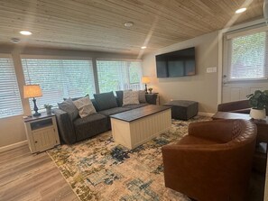 Ein Wohnzimmer mit einer Sofagarnitur, einem Couchtisch, einem braunen Sessel, einem Beistelltisch mit Lampe und einem an der Wand montierten Fernseher.