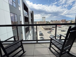 A balcony with two chairs, a view of a parking lot, and a cityscape with buildings in the background.