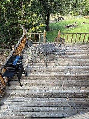 Eine Holzterrasse mit einem Metalltisch und -stühlen, einem Grill und Blick auf eine grüne Fläche mit Bäumen.