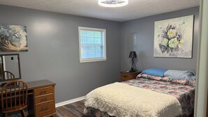 Un dormitorio con cama, escritorio, silla, ventana y obras de arte en las paredes.