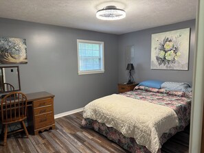 Un dormitorio con cama, escritorio, silla, ventana y obras de arte en las paredes.