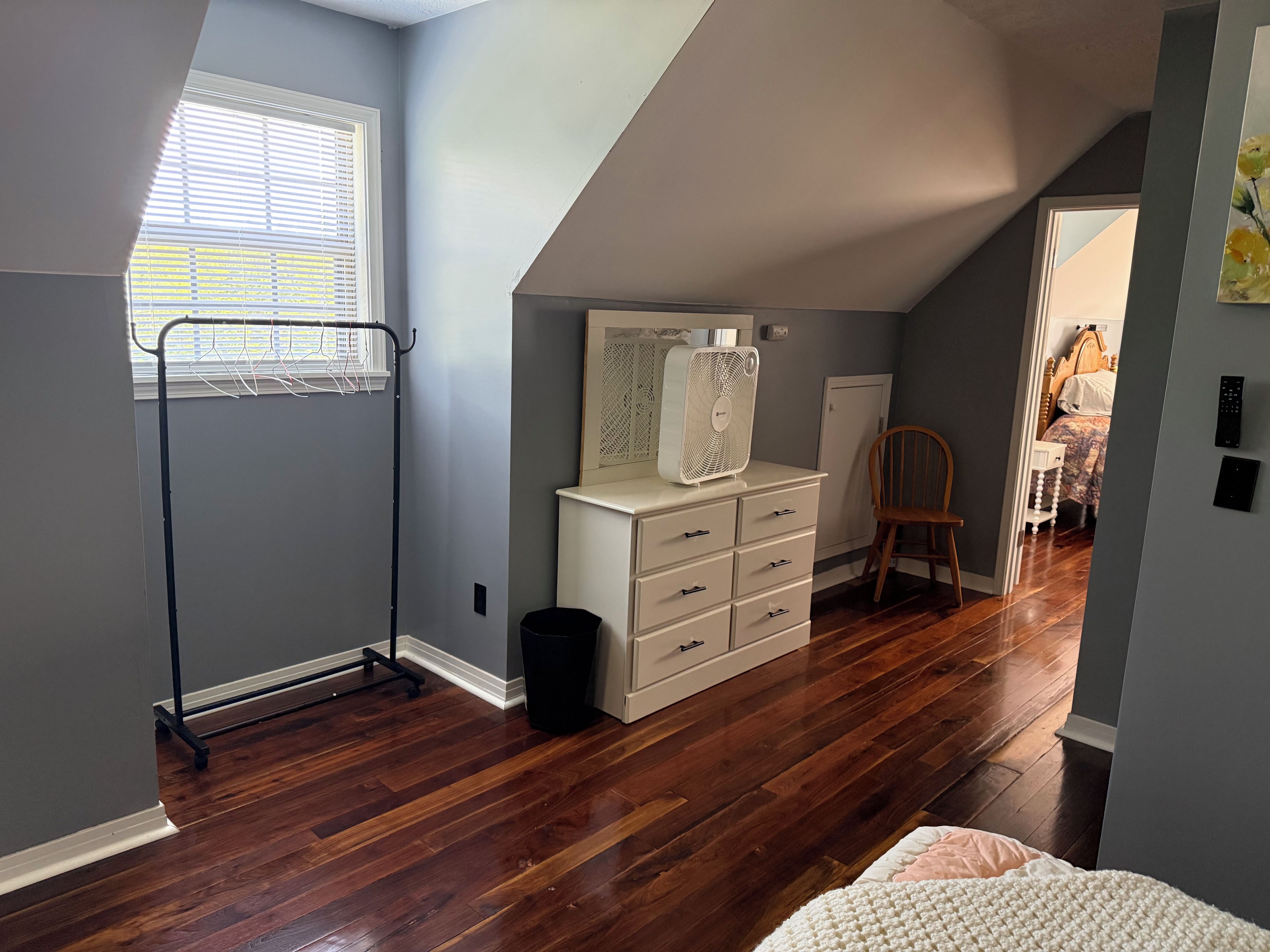 A room with a sloped ceiling, a white dresser, a wooden chair, and a window with blinds.